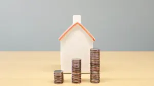 Mažas namo modelis ir krūvelės monetų ant stalo simbolizuoja būsto paskolos palūkanas.