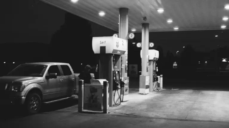 Tamsus kadras, kuriame automobilis stovi degalinėje vėlai vakare, svarstant apie kuro kainas.