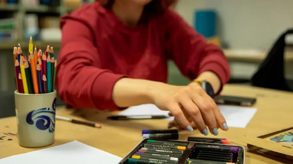 Mergina raudonu džemperiu sėdi prie stalo ir ruošiasi piešti spalvotais aliejiniais pastelėmis.