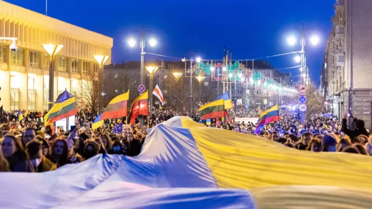 Didelė minia žmonių laiko Ukrainos vėliavą gatvėje su Lietuvos vėliavomis vakaro metu.
