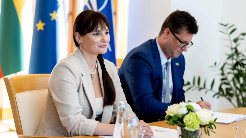 Moteris ir vyras sėdi prie stalo su Europos Sąjungos ir NATO vėliavomis fone.