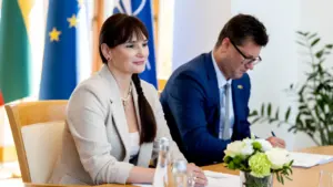 Moteris ir vyras sėdi prie stalo su Europos Sąjungos ir NATO vėliavomis fone.
