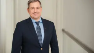 Vidutinio amžiaus vyras su tamsiu švarku, melsvais marškiniais ir dryžuotu kaklaraiščiu šypsosi žiūrėdamas į kamerą.