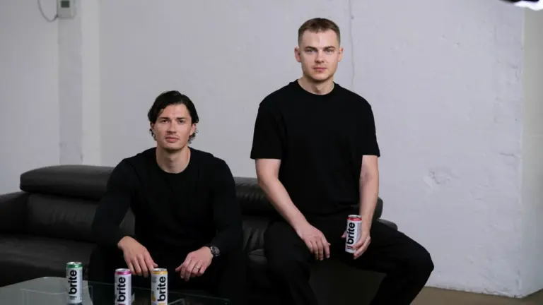 Du jauni vyrai vilki juodus drabužius ir pozuoja šalia „Brite“ gėrimų skardinių patalpoje.
