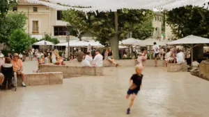 Saulėta miesto aikštė su žmonėmis, poilsio zona ir bėgančiu vaiku Marijampolės parko nuotaikai.