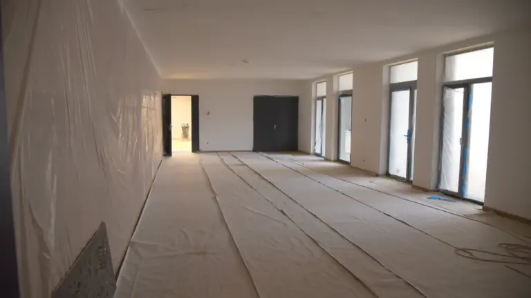 Didelė tuščia patalpa su baltomis sienomis, uždengtomis grindimis ir dideliais langais statybų metu.