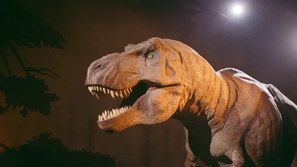 Didelio dinozauro maketas su atvira burna, apšviestas teatrinio stiliaus scenos šviesomis.