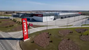 Didelis „Rimi“ logistikos centras su raudonu stulpu ir Lietuvos vėliava saulėtą dieną.