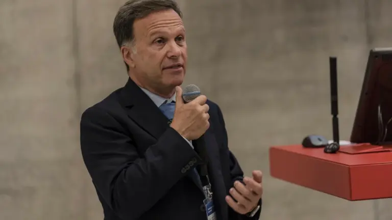Vidutinio amžiaus vyras su kostiumu laiko mikrofoną ir kalba konferencijoje, stovėdamas prie raudonos pakylos.
