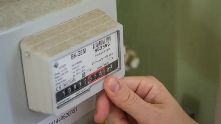 Žmogaus ranka tikrina namų elektros ar dujų skaitiklio rodmenis, aiškus vaizdas.