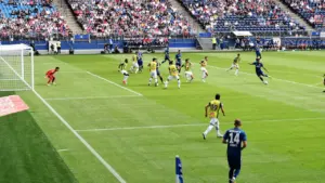 Profesionali futbolo rungtynių akimirka dideliame stadione su žaidėjais aikštėje ir žiūrovais tribūnose.