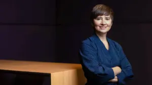 Trumpų plaukų moteris su tamsiai mėlynos spalvos dryžuotu švarku stovi biure, sukryžiavusi rankas ant krūtinės.