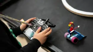 Mokinys laiko nuotolinio valdymo pultelį ir valdo mažą savadarbį robotą ant juodo stalo.