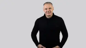 Vidutinio amžiaus vyras trumpais plaukais, vilkintis juodą megztinį, stovi šviesiame fone rankas įsidėjęs į kišenes.